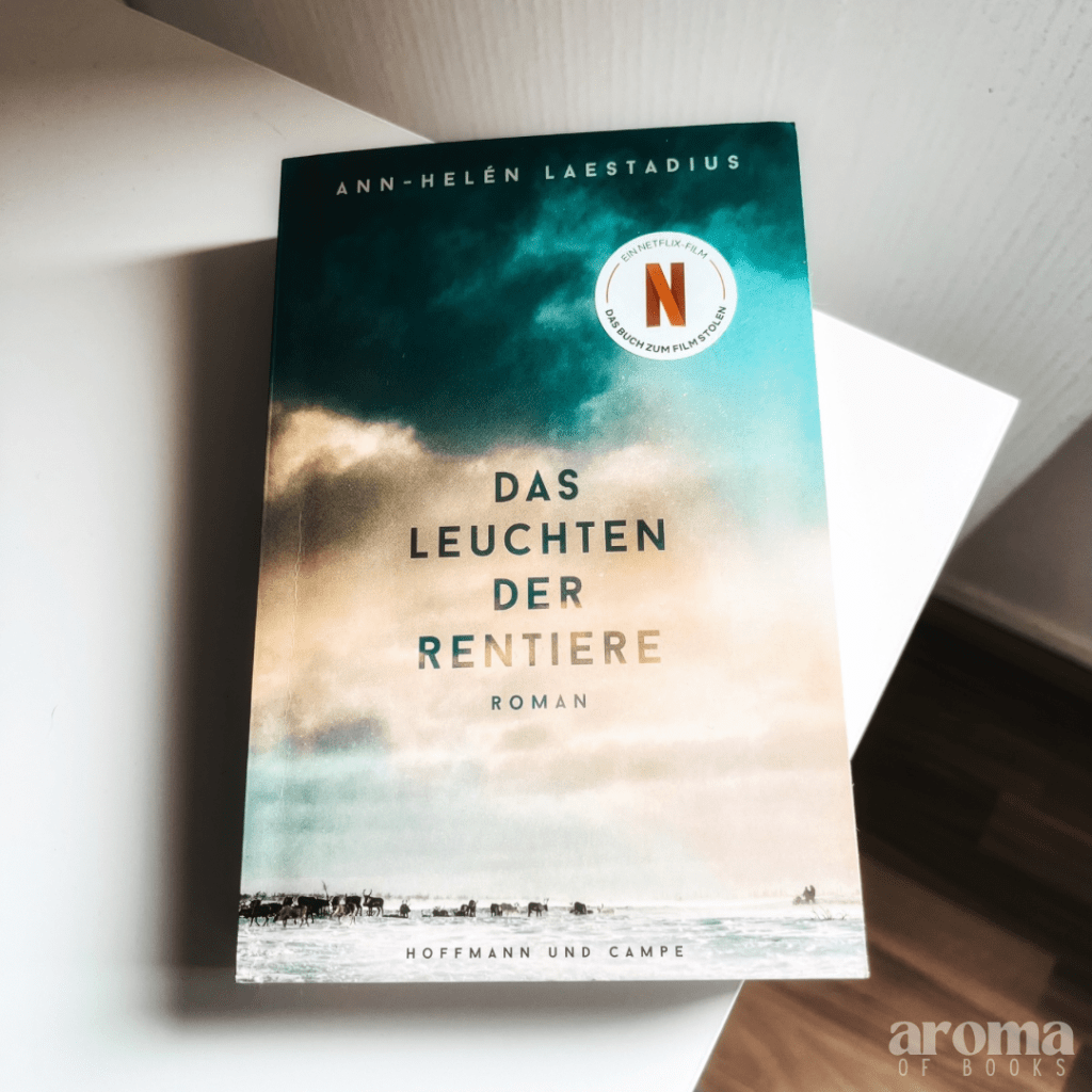 Man sieht das Buch auf einer weißen Kommode liegen. Auf dem Cover ist eine Schneelandschaft und ein wolkiger Himmel. In weiter Entfernung sind Rentiere zu sehen.